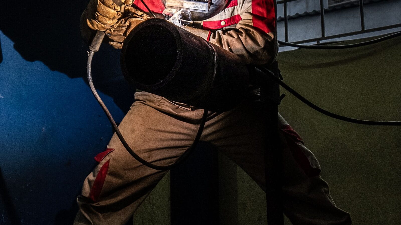 Welder in protective equipment welding a steel pipe at night, sparks lighting the joint