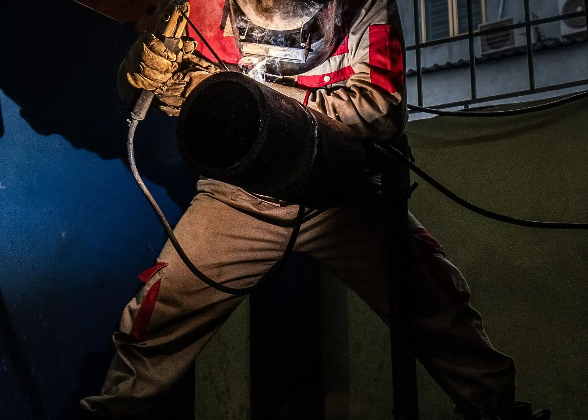 Welder in protective equipment welding a steel pipe at night, sparks lighting the joint