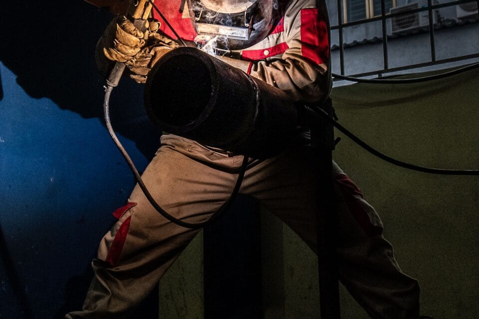 Welder in protective equipment welding a steel pipe at night, sparks lighting the joint
